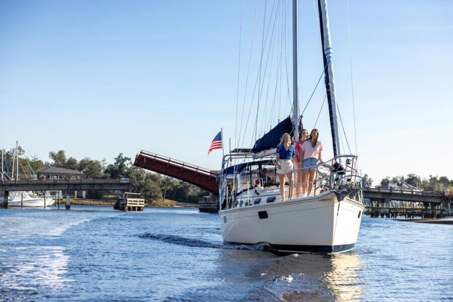 Panama City’s Multi-Coastal Story: History, Culture & Life on the Bay - 5 A sailboat with three people on the bow passes under an open drawbridge on a sunny day in Panama City FL, with an American flag on the stern and a marina in the background—a perfect travel moment beyond the beach.