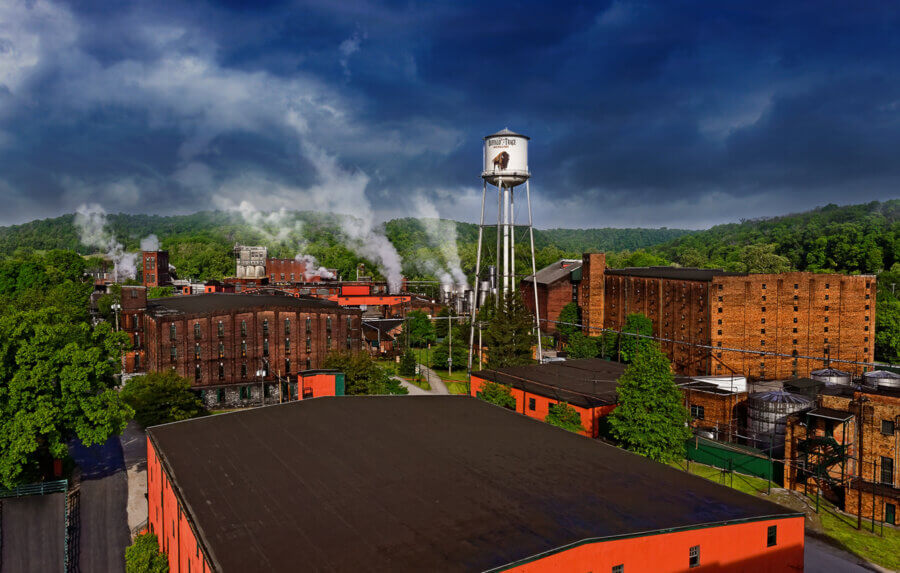 Are These Kentucky's Most Beautiful Distilleries? - 2 Industrial complex with multiple brick buildings, a tall white water tower, and steam rising set against dense green trees under a dramatic cloudy sky.