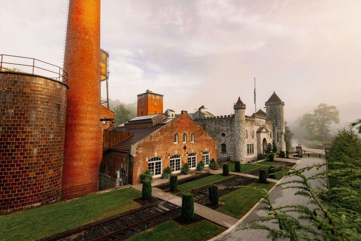 A historic Southern distillery with brick and stone buildings, tall smokestacks, and visible train tracks sits surrounded by greenery on a misty day—a timeless setting to explore the legacy of Southern whiskey.