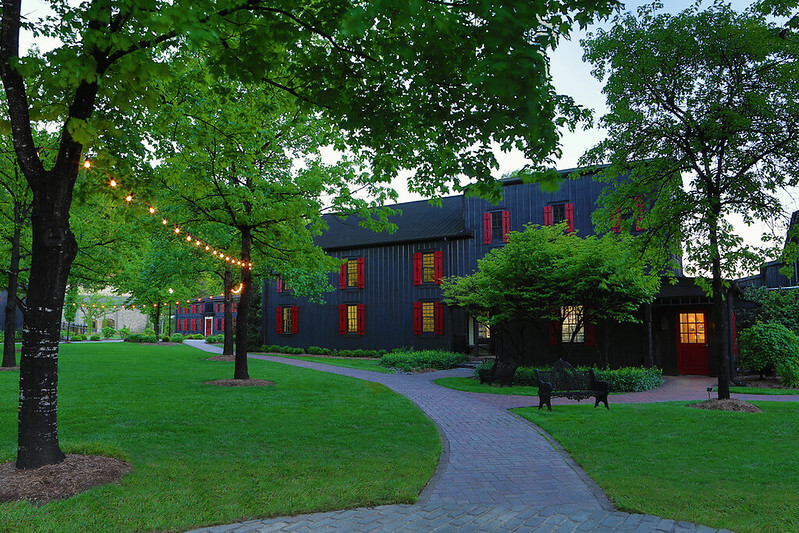 Are These Kentucky's Most Beautiful Distilleries? - 10 A brick pathway leads to a black building with red windows, surrounded by green lawns and trees. String lights hang between the trees, inviting you to explore distilleries on scenic Kentucky distillery tours.