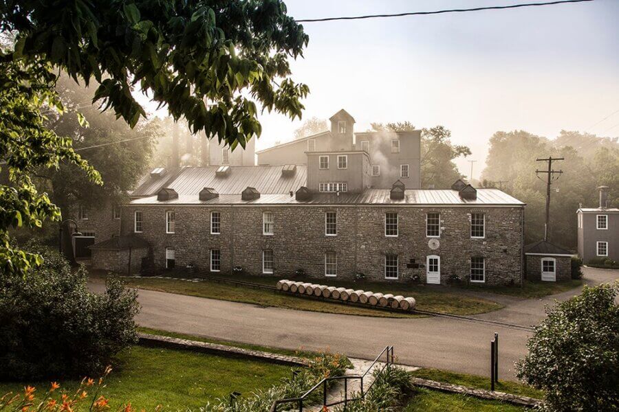 Are These Kentucky's Most Beautiful Distilleries? - 13 Historic stone distillery building with a metal roof, smoke rising from vents, surrounded by greenery and a paved road in front.