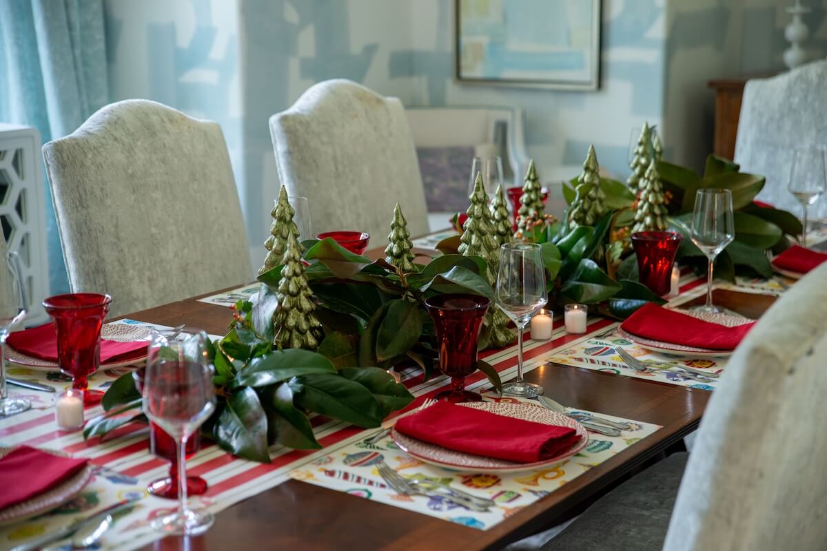 A dining table set for a holiday meal with red napkins, red glasses, greenery, decorative Christmas trees, and candles as the centerpiece—perfect inspiration for holiday hosting or table-setting tips from an event planner.