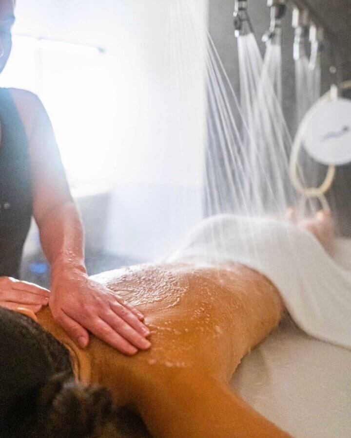 15 Decadent Nashville Spa Treatments to Gift - 2 A person enjoys a relaxing back massage as water streams from overhead showers in a spa setting, offering the same rejuvenation as popular Nashville facials.