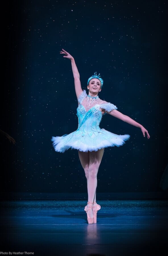 Meet the Star of Nashville Ballet’s Nutcracker - 3 Colette Tilinski, in a white tutu and tiara, dances en pointe on stage with one arm raised against a dark, starry background.