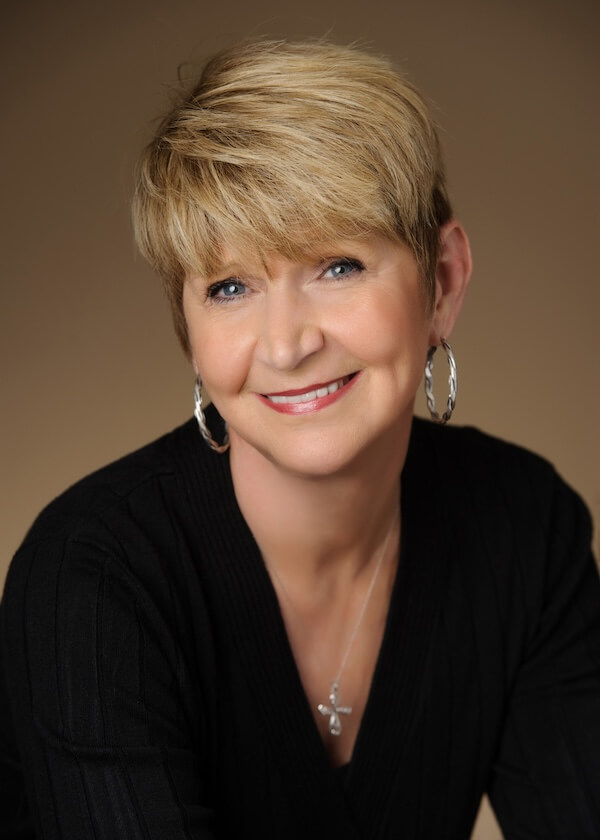 From Kentucky Bourbon to Cancer Advocacy: This Renaissance Woman Does It All - 2 Smiling woman with short blonde hair wearing hoop earrings, a black top, and a cross necklace, posed against a plain brown background reminiscent of Burnt Tavern Bourbon’s rich, warm hues.
