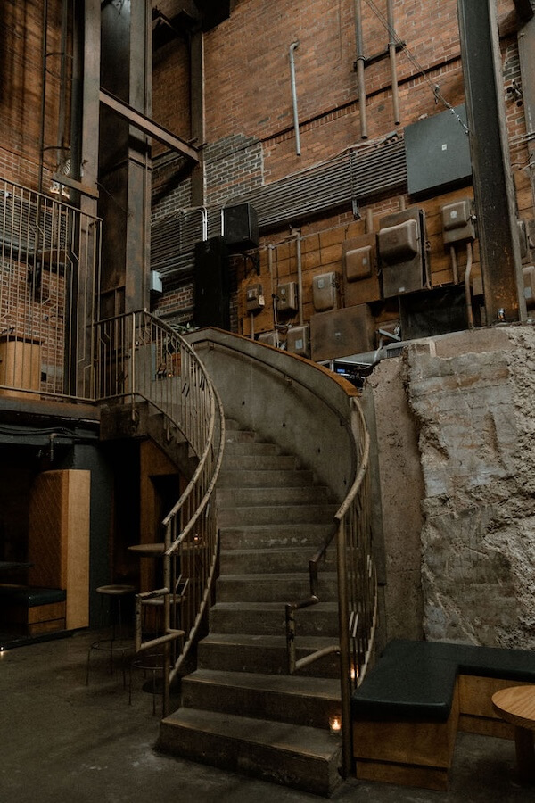 Meet the Woman Behind Old Glory & ELEVEN11 - 3 A curved concrete staircase with a metal railing, designed by Alexis Soler, leads to an upper level in an industrial-style interior with exposed brick walls and electrical panels.