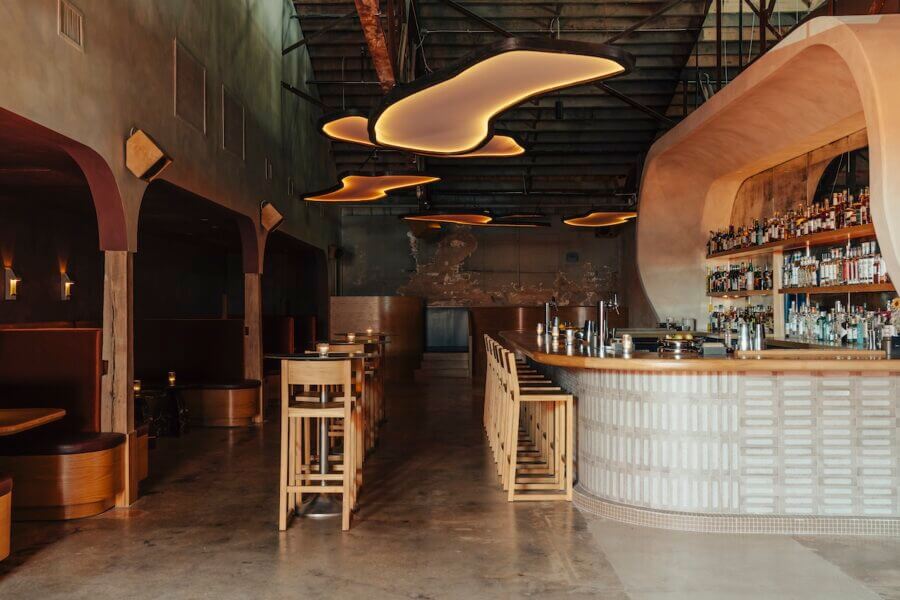 Meet the Woman Behind Old Glory & ELEVEN11 - 4 Modern bar interior designed by Alexis Soler, featuring a curved wooden bar, high stools, booth seating, exposed ceiling, and abstract ceiling lights. Shelves behind the bar are stocked with bottles. Space is empty.
