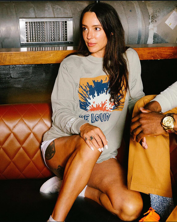 Meet the Woman Behind Old Glory & ELEVEN11 - 6 Alexis Soler, with long dark hair, sits on a tan quilted bench wearing a graphic sweatshirt and white shorts, one leg crossed to reveal a tattoo on her thigh.