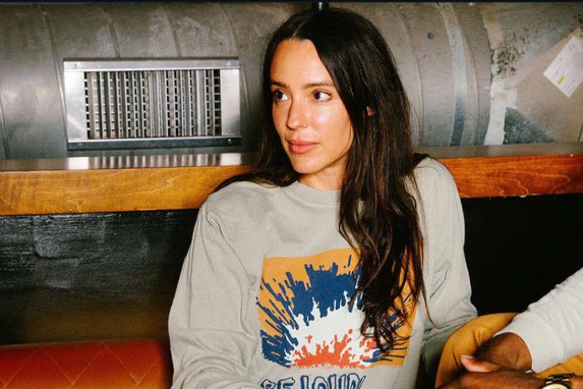 A woman with long dark hair sits indoors on a bench, wearing a light-colored sweatshirt with a graphic print, looking to the side, reminiscent of Alexis Soler's relaxed style.