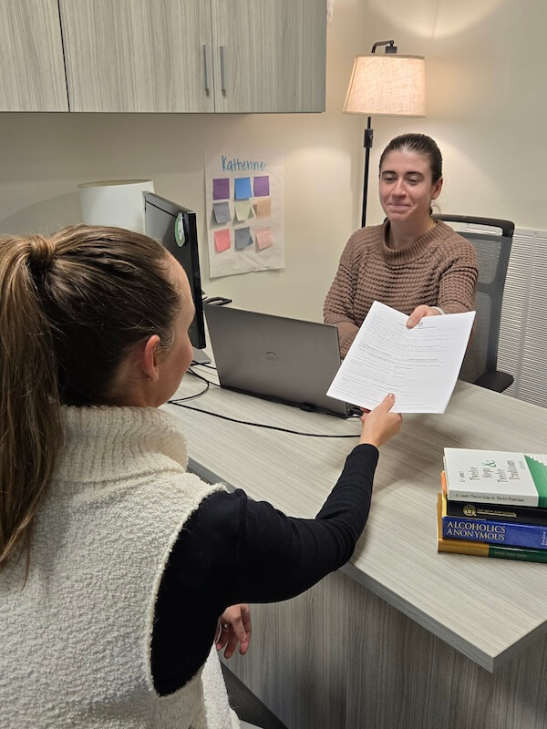 This Nashville Mom Rebuilt Her Life at Renewal House - 2 Two women sit across a desk in a Renewal House office. One hands paperwork to the other, who is seated by a laptop. A lamp, books, and a wall chart are visible in the background.