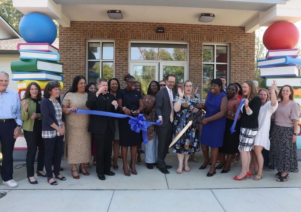 A group of people, including families, stands in front of Renewal House, gathered around a blue ribbon for a ribbon-cutting ceremony. Large book-themed columns frame the entrance.