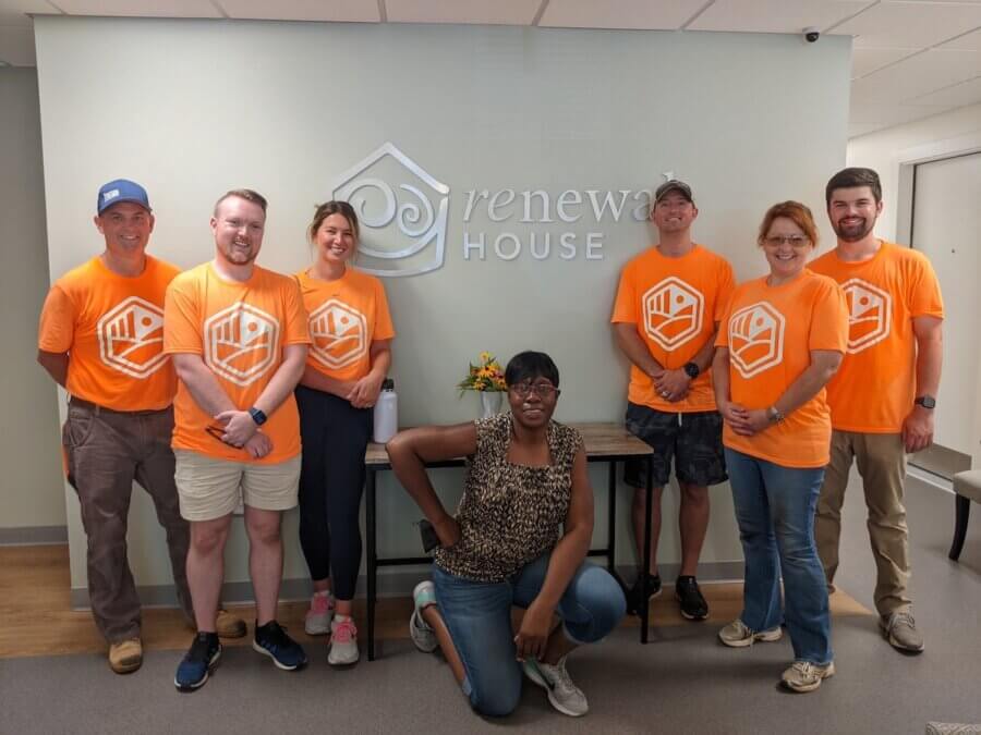 Renewal House: Where Mothers Heal & Families Begin Again - 5 Seven people in matching orange t-shirts stand and kneel indoors in front of a wall displaying the Renewal House sign.