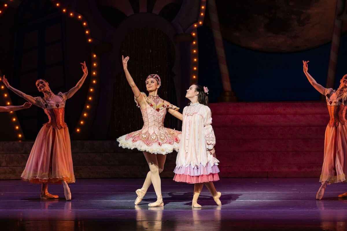 Ballet dancers perform on stage; Colette Tilinski, in a tutu, poses en pointe while another in a nightgown stands beside her. Two other dancers are in the background with arms raised.