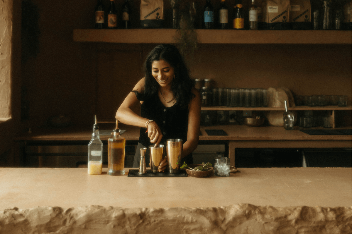 Shivani Darsinos stands behind the bar counter at Cafe Babu, preparing a drink amid neatly arranged bottles, utensils, and ingredients. Shelves lined with bottles and glasses fill the inviting background.