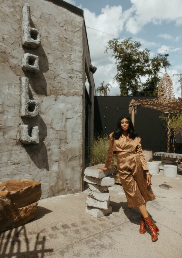 She's Behind Nashville's Dreamiest Coffee Shop - 2 Shivani Darsinos, wearing a tan outfit and red boots, leans against a stone table outside cafe babu, with its name displayed on the wall behind her.