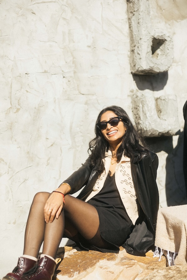 She's Behind Nashville's Dreamiest Coffee Shop - 5 Shivani Darsinos, with long dark hair, wearing sunglasses, a beige vest, black dress, and boots, sits on a stone bench against a textured wall outside cafe babu, smiling at the camera.