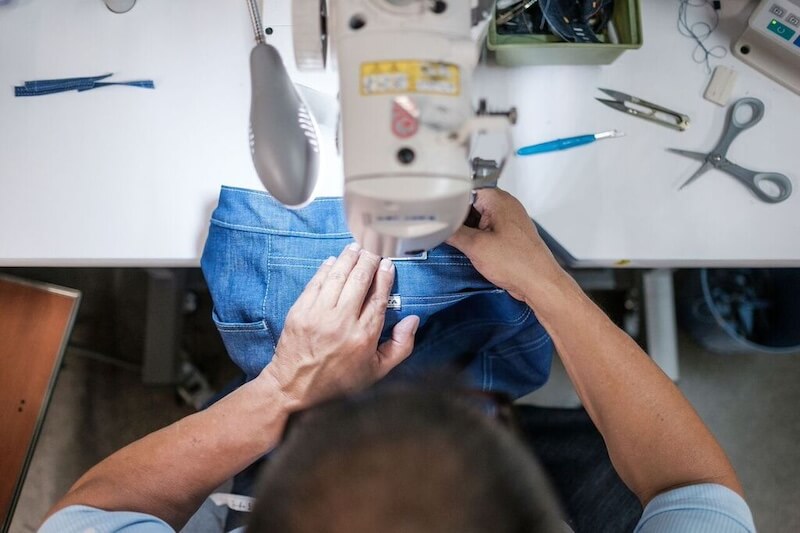 Blue Delta Jeans: Made in MS, One Pair at a Time - 6 Person sewing blue denim jeans with a sewing machine at a worktable; scissors, thread, and sewing tools are nearby, crafting Blue Delta Jeans proudly Made in Mississippi.