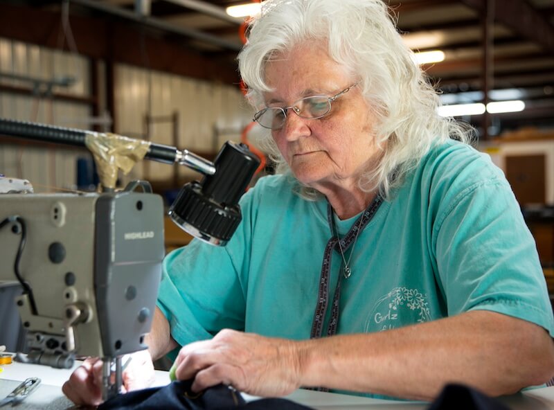 Blue Delta Jeans: Made in MS, One Pair at a Time - 3 An older woman with white hair and glasses operates an industrial sewing machine in a Mississippi workshop, focusing on fabric under the needle—crafting premium Blue Delta Jeans.