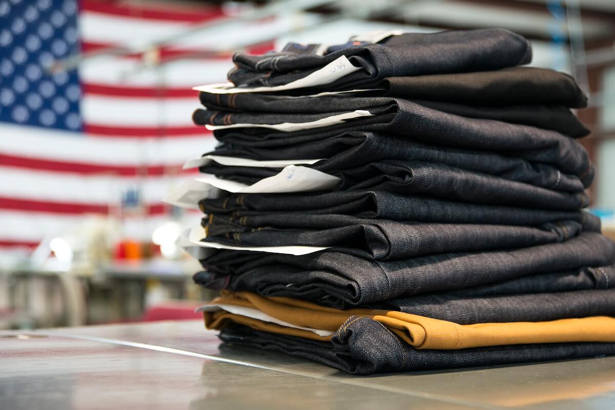 A stack of folded jeans on a table, with an out-of-focus American flag in the background.
