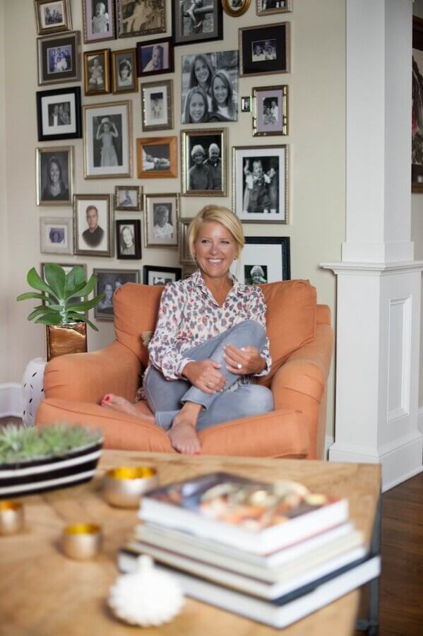 Happy Everything! Laura Johnson’s Joyful Approach to Home & Hosting - 2 A woman sits barefoot on an orange armchair, smiling, in a living room with a wall covered in framed family photos. A coffee table with books and decor is in the foreground.