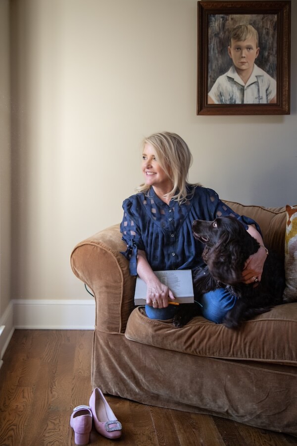 Happy Everything! Laura Johnson’s Joyful Approach to Home & Hosting - 7 Laura Johnson sits on a brown couch holding a book and her black dog, with pink shoes on the floor and a framed painting of a boy behind her, embodying the spirit of Happy Everything home hosting.