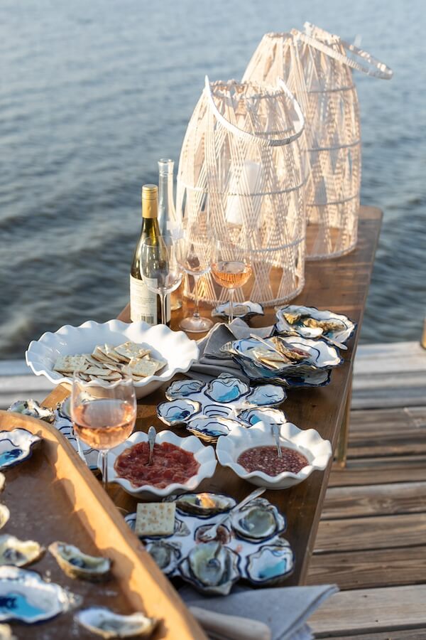 Happy Everything! Laura Johnson’s Joyful Approach to Home & Hosting - 12 A wooden table by the water displays oysters, crackers, spreads, wine, glasses, and two wicker lanterns—set for an outdoor meal inspired by Happy Everything and styled beautifully by Laura Johnson.