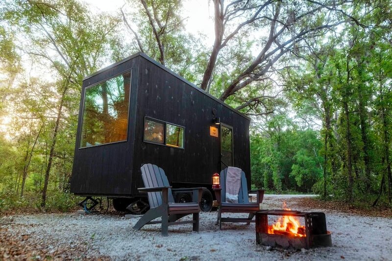 10 Digital Detox Destinations Around the South - 4 A small black tiny house on wheels is parked in a wooded area with two chairs and a fire pit in front, making it one of the best places for a peaceful digital detox.