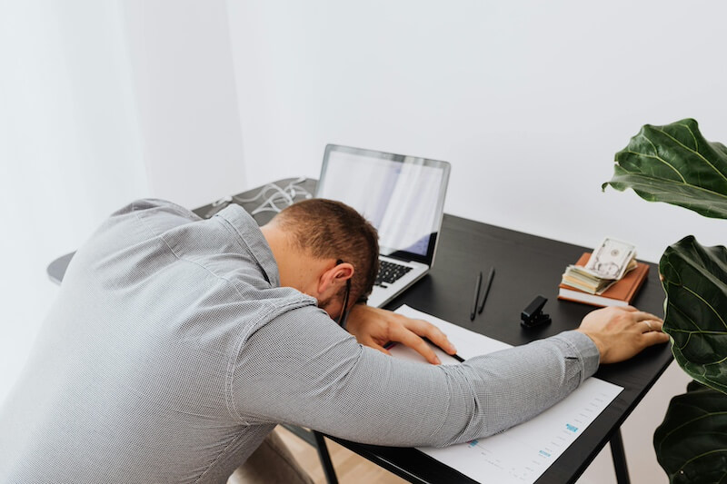 Grammar Guru: 12 Clever "Britishisms" to Adopt - 3 A person sits at a desk with their head down on paperwork, next to a laptop, phone, glasses, and a stack of cash—perhaps pondering which Britishisms to adopt during their busy day.