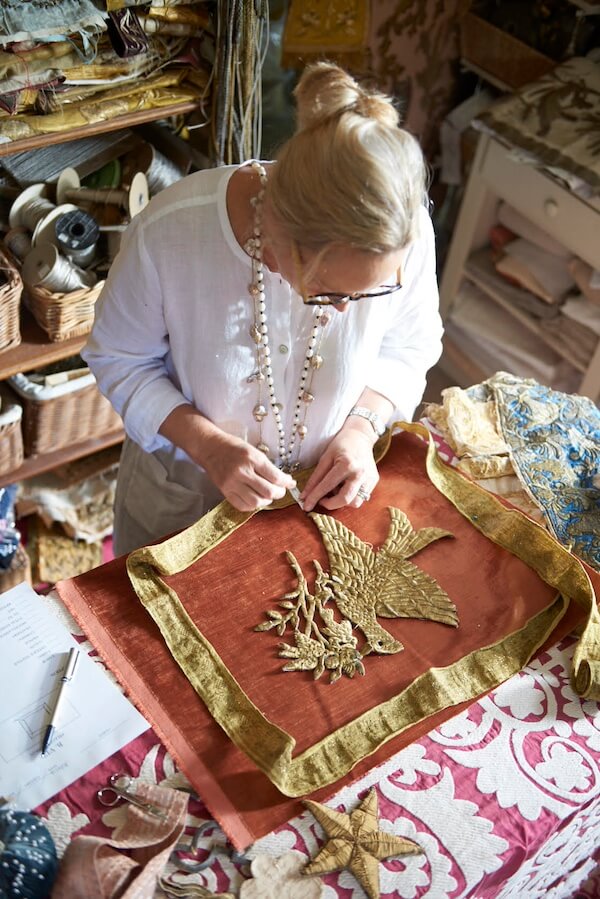 Pillow Talk with B. Viz Design's Rebecca Vizard - 8 A person in a white shirt works on an embroidered textile with a gold bird design at a table covered with various fabrics and sewing materials.
