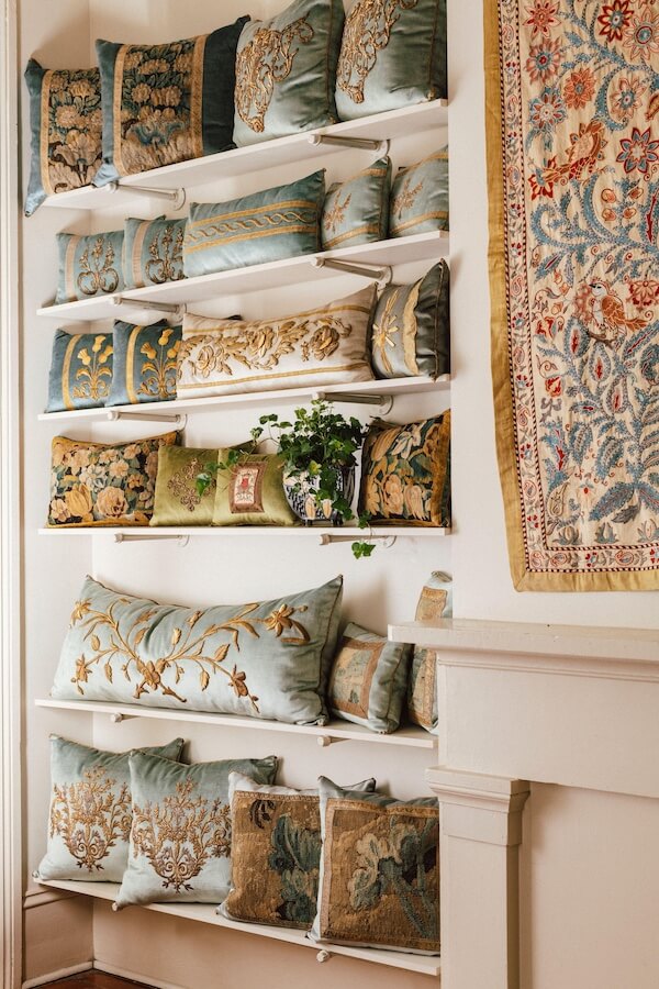 Pillow Talk with B. Viz Design's Rebecca Vizard - 6 Four shelves display decorative pillows with intricate floral embroidery in blue, gold, and green tones—a striking example of viz design; a potted plant and a tapestry hang nearby.