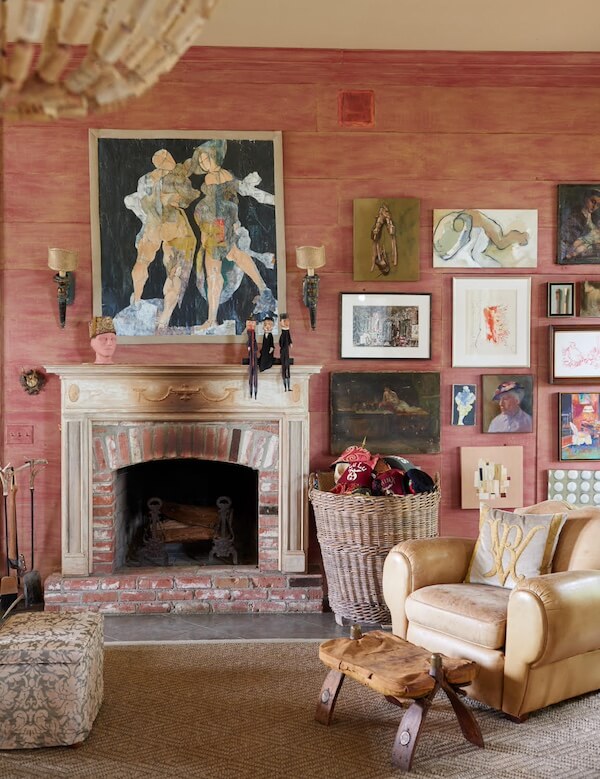 Pillow Talk with B. Viz Design's Rebecca Vizard - 5 A living room designed with b. viz design flair features a pink textured wall, brick fireplace, eclectic artwork, a leather armchair, rustic wooden table, and a large basket filled with cozy blankets.