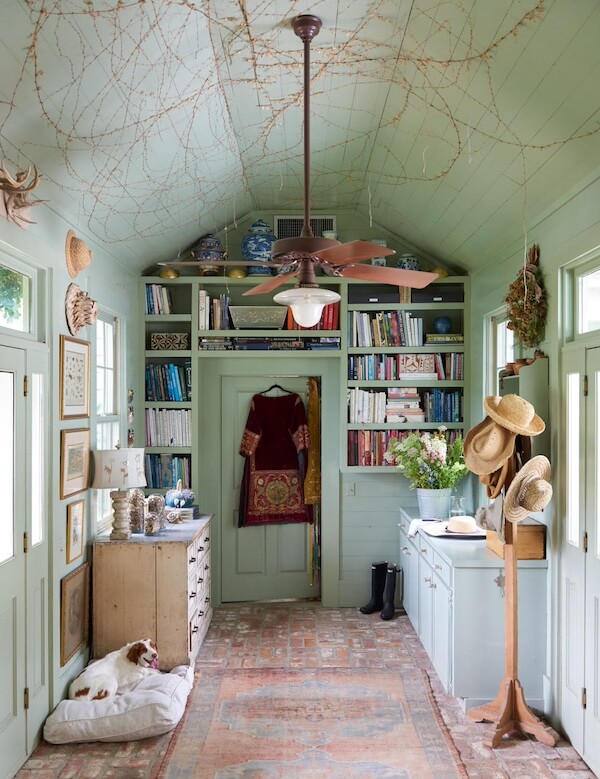 Pillow Talk with B. Viz Design's Rebecca Vizard - 4 A narrow room with shelves of books, decorative jars, a hanging dress, a ceiling fan, hats on hooks, flowers, boots, and a dog resting on a bed atop the patterned rug.