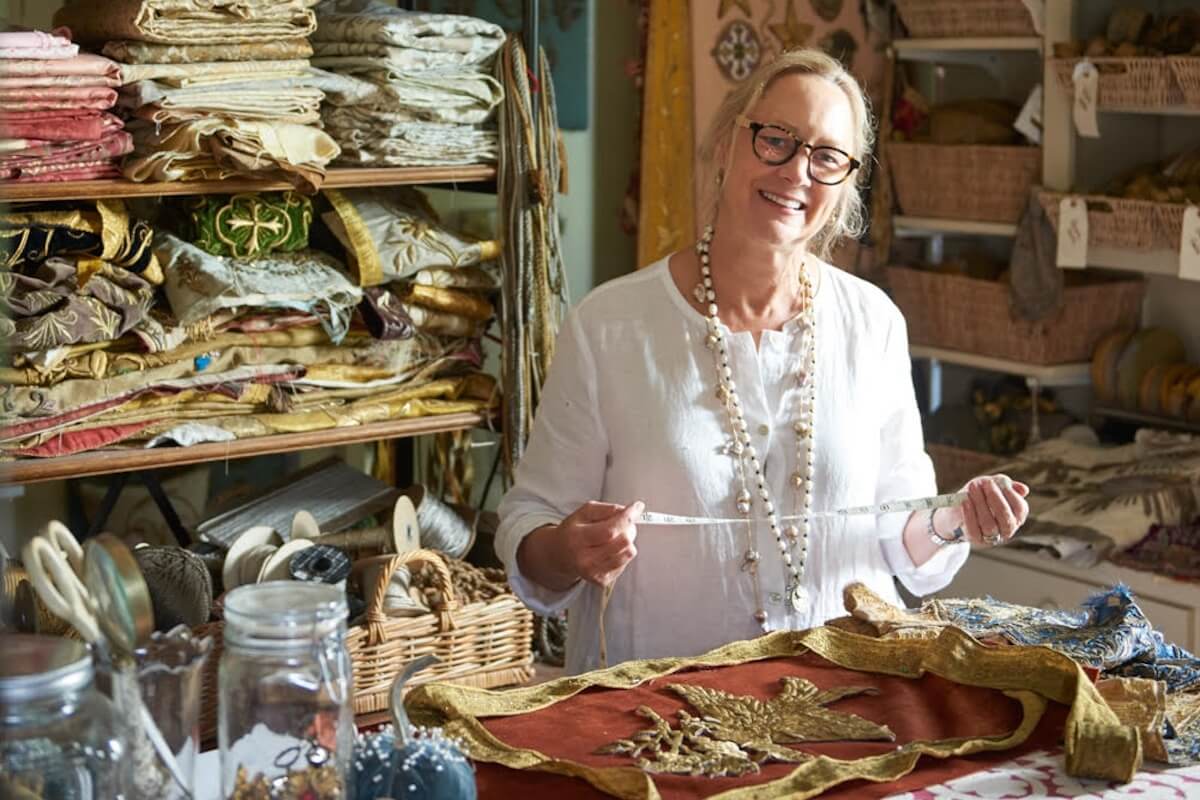 A woman wearing glasses and a white blouse smiles while measuring fabric amidst various textiles and sewing supplies, creating a scene that reflects her passion for viz design.