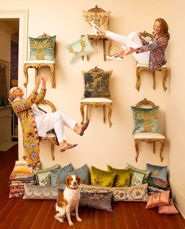 Pillow Talk with B. Viz Design's Rebecca Vizard - 2 Two women pose playfully on vintage chairs mounted on a wall, styled with decorative pillows and b. viz design touches. A dog relaxes among stacked pillows, completing the creative scene.