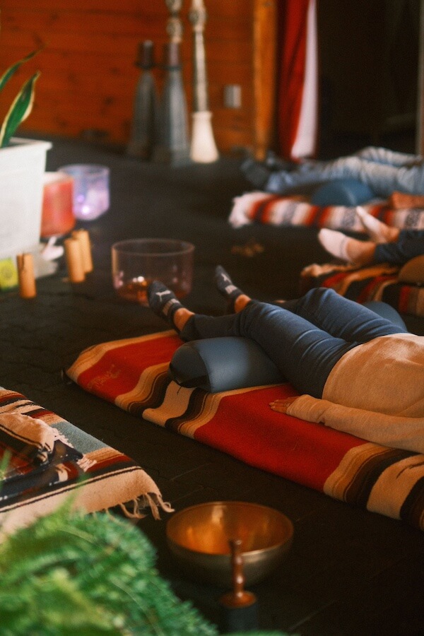 10 Digital Detox Destinations Around the South - 5 People lying on mats with pillows under their legs in a cozy room, surrounded by sound bowls and blankets, enjoying a digital detox session focused on relaxation or meditation.
