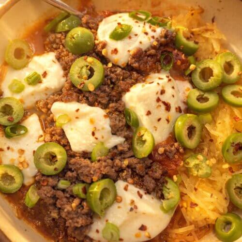 My Go-To Spaghetti Squash "Pasta" Bake - 3 A bowl of Spaghetti squash pasta bake topped with ground beef, melted cheese, green olives, scallions, and red pepper flakes—a healthy recipe with a fork on the side.