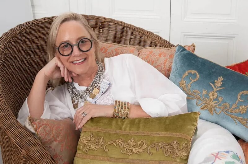 Pillow Talk with B. Viz Design's Rebecca Vizard - 3 A woman with round glasses and layered necklaces lounges on a wicker chair with patterned pillows, smiling at the camera.