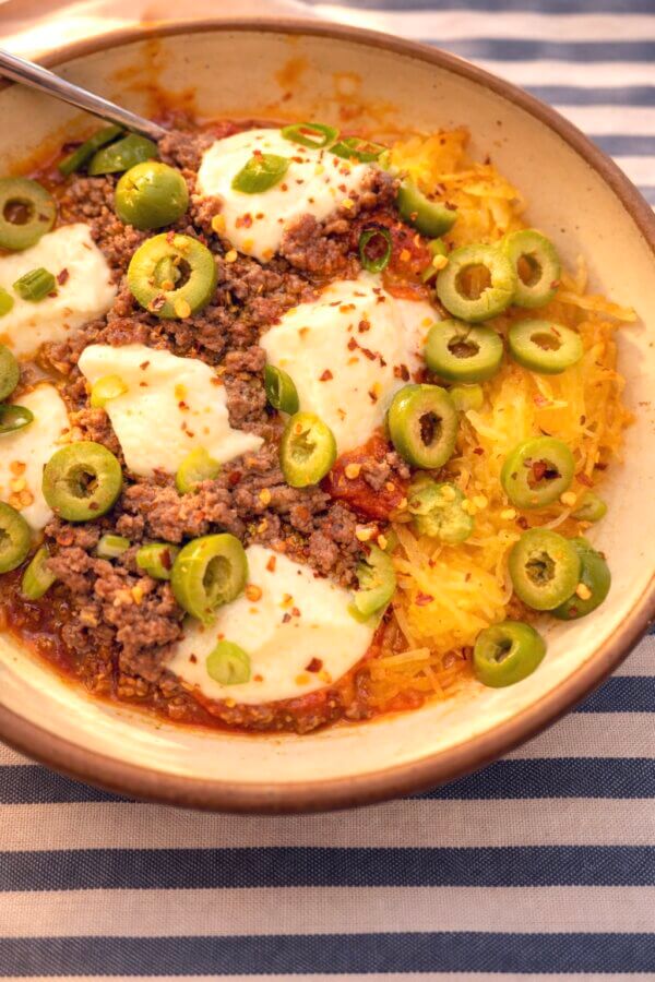 My Go-To Spaghetti Squash "Pasta" Bake - 2 A bowl of spaghetti squash pasta bake topped with ground meat, tomato sauce, melted cheese, green olives, green onions, and red pepper flakes makes for a healthy recipe. A spoon rests inside the bowl for easy serving.