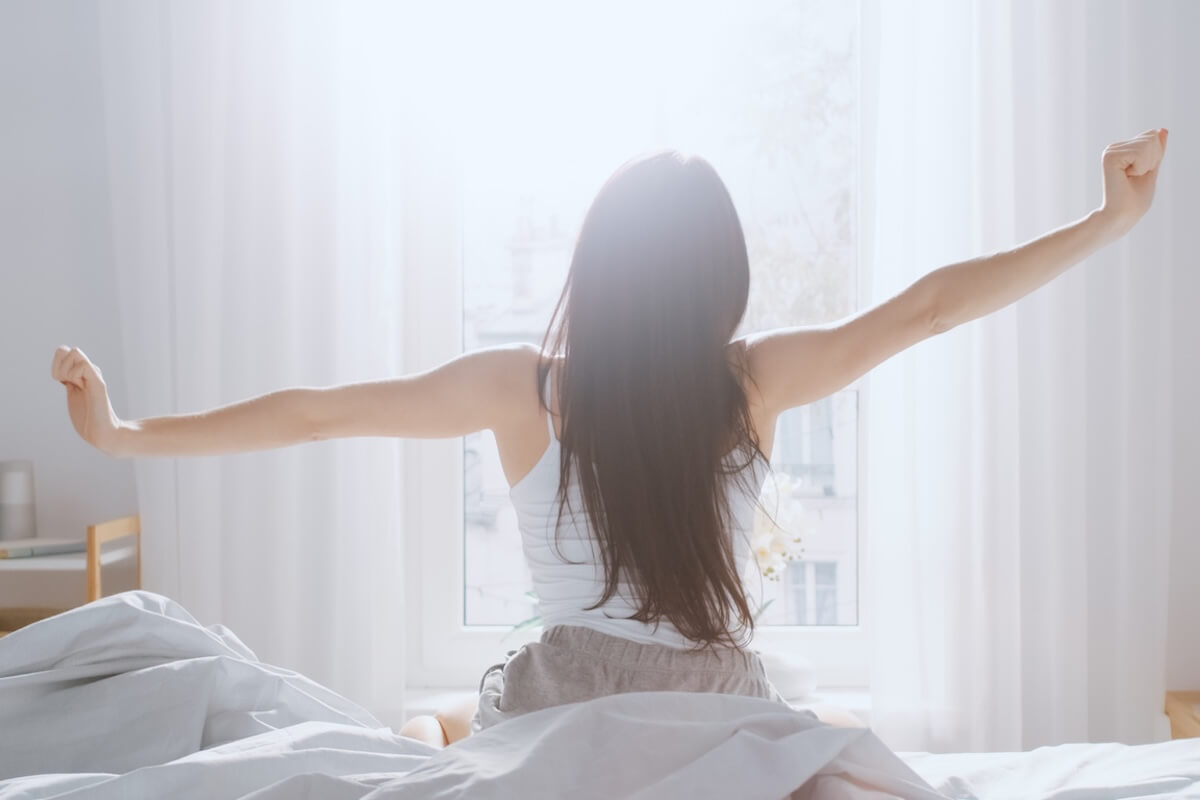 A person with long hair stretches their arms while sitting on a bed facing a bright window with white curtains, capturing the essence of 2026 trends and what's in and out for serene morning routines.