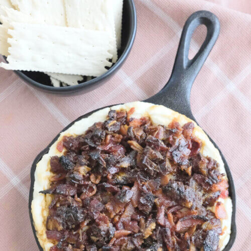 RECIPE: Hot Honey Goat Cheese Dip with Bacon and Dates - 2 A small cast iron skillet filled with a baked dip topped with chopped bacon and dates, next to a bowl of rectangular crackers on a pink plaid cloth—perfect inspiration for anyone searching for easy appetizer recipes.