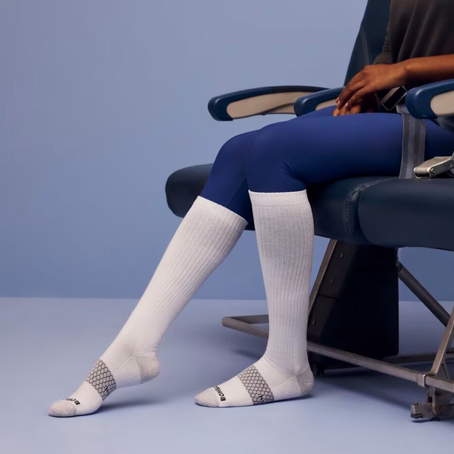 Buy It for Life: 5 Long-Lasting Travel Essentials Loved by Our Readers - 4 A person wearing blue leggings and white compression socks sits in an airplane seat against a light blue background.