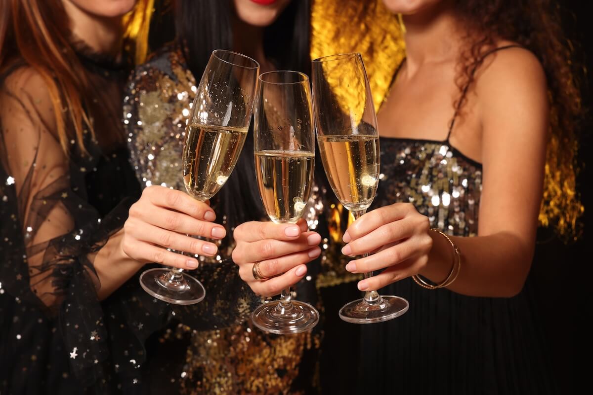 Three women in sparkly dresses hold and clink champagne glasses together at a lively New Year's Eve Birmingham celebration.