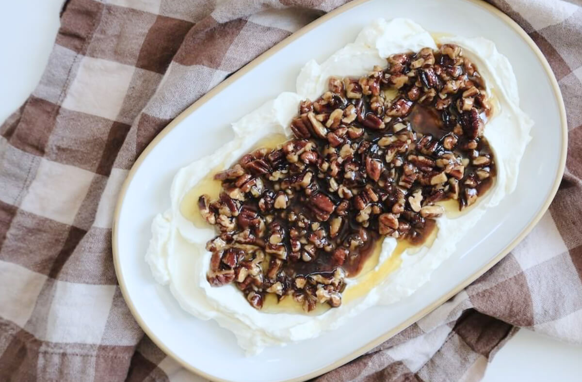 A serving platter with a layer of whipped cream cheese topped with chopped pecans and honey sits on a brown and white checkered cloth—perfect for easy appetizer recipes.