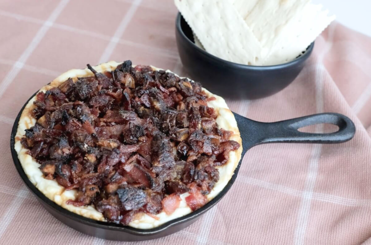 A skillet pie topped with cooked, diced meat sits on a pink plaid cloth next to a black bowl filled with flatbread and creamy goat cheese dip.