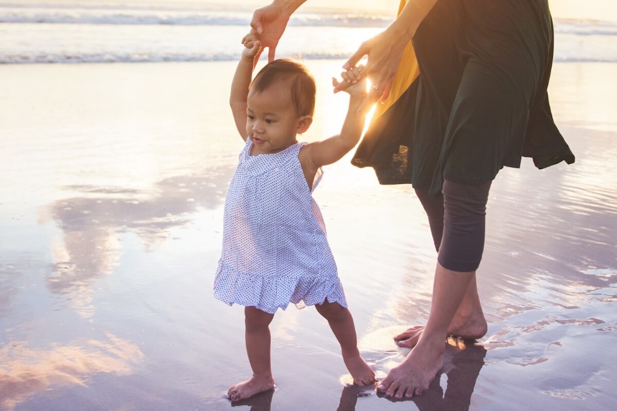 A toddler in a white dress walks on a wet sandy beach, holding hands with an adult as the sun sets in the background, capturing a heartwarming moment reminiscent of Renewal House’s spirit of connection and hope.