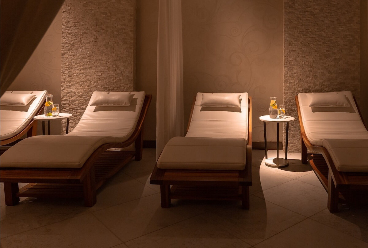 Three cushioned lounge chairs with pillows are arranged in a dimly lit room, each beside a small round table holding a glass of drink and a bottle—perfect for relaxing after indulgent Nashville facials.