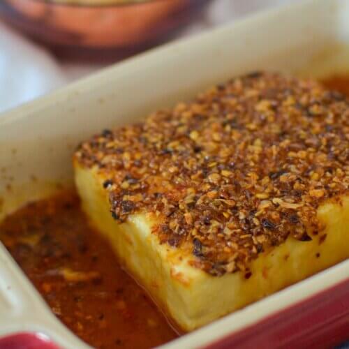 RECIPE: Spicy Honey Baked Feta Dip - 2 A block of baked feta cheese topped with herbs and spices sits in a rectangular baking dish with some sauce around it.