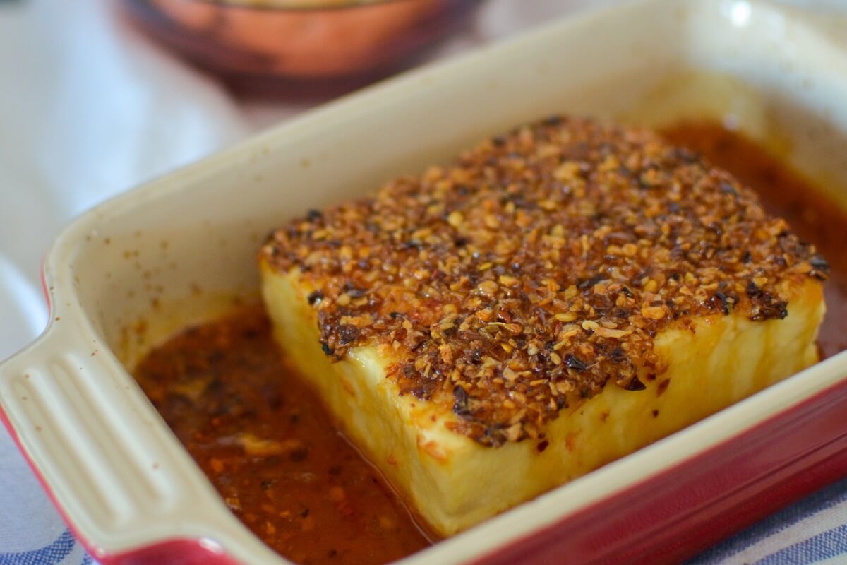 A block of baked feta cheese topped with herbs and spices sits in a rectangular baking dish with some sauce around it.
