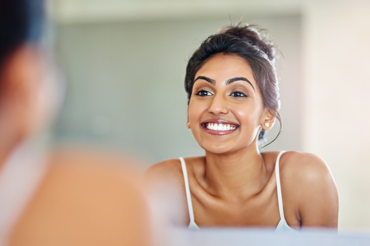 A young woman with dark hair in a bun, wearing a white tank top, smiling broadly after discovering teeth whitening myths debunked while looking at her reflection in a mirror.