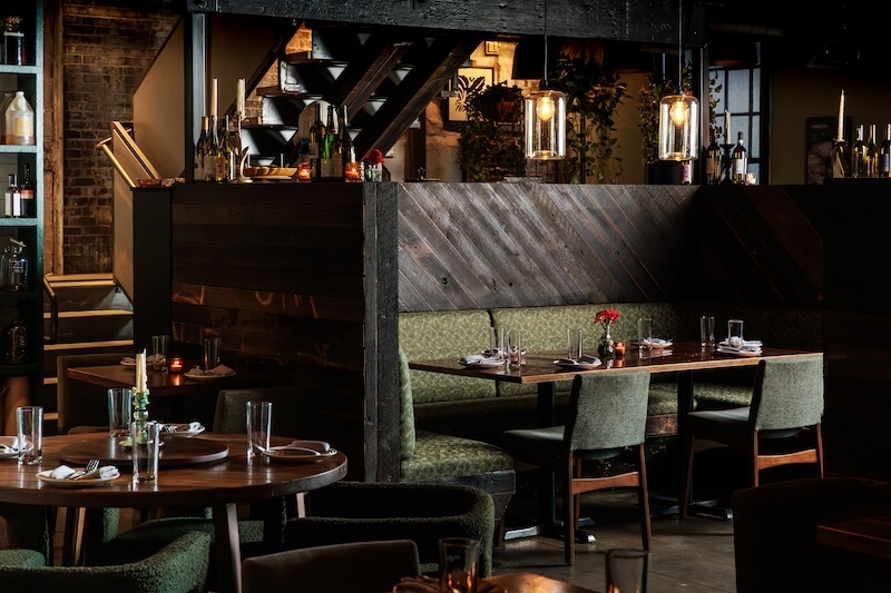 A dimly lit restaurant interior with empty tables set with plates, glasses, and napkins, featuring dark wood decor and green upholstered chairs—an inviting scene among the new Nashville restaurants.